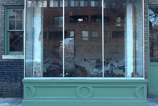 Photograph of a glass picture-window of a storefront, in which the glass reveals a view of demolition happening inside the storefront, while it also reflects the image of the exterior of a building across the street. To the left and right side of this large picture-window are smaller windows. The window to the left reflects or reveals nothing, while the window to the right reflects some light.