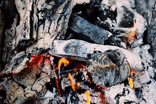 A bundle of wood charcoal, covered in white ash and orange hued flames running throughout