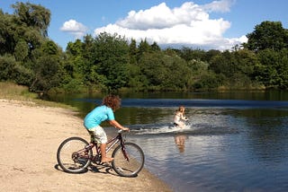 2 children in swimwear bike into a lake, one is poised at the edge of the water, the other’s bike is already submerged