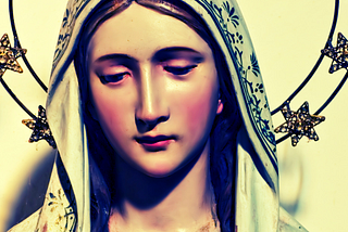A porcelain Madonna statue with rosy red cheeks, surrounded by a halo of stars. Dominant colors of tawny yellow, green, pink, and blue shadow.