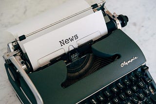 A black typewriter with a sheet of paper in it that has the word, “News” typed on it.
