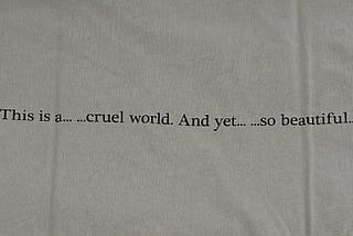 “This is a… …cruel world. And yet… …so beautiful…” printed on the back of a grey cotton tee shirt.