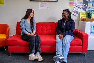 Two people sit smiling on a red sofa, both are wearing casual clothes.