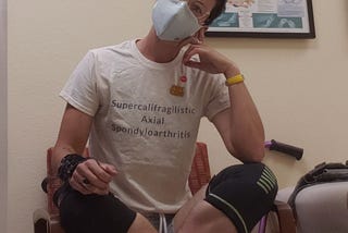 A portrait of me sitting cross-legged in a chair in a clinical exam room. I’m white-appearing with short dark hair. I’m wearing glasses, a light blue face mask over an N95, a t-shirt that says, “supercalifragilistic axial spondyloarthritis,” shorts and compression knee sleeves.