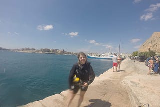 Emily on the dock of a bay in Malta, decked out in all her scuba diving gear with a big smile on her face