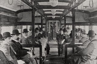 The interior of a train carriage filled with passengers, most of whom are men wearing suits and bowler hats. Despite the scene clearly being old-fashioned it is also reminscient of a modern train.