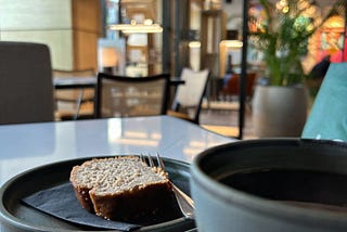 In a cafe. A close up of a dark plate with a slice of banana loaf, and the edge of a coffee cup.
