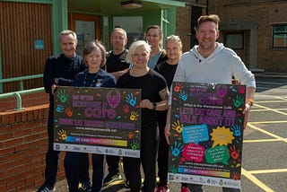 Seven people, men and women, standing together in front of a building at MDP Headquarters and those standing at the front are holding large posters advertising the Menopause Cafe