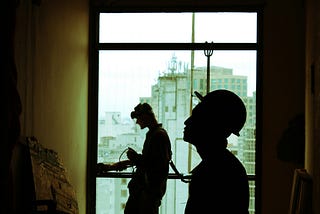 Two construction workers in shadow before a window. Improvement is ongoing.