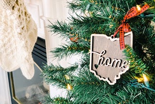 A Christmas tree in front of a fireplace with a wooden ornament in the shape of a state with the word “home,” written on it