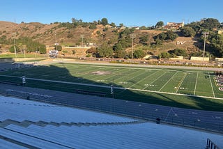 A photo of the inside of Whittier College’s football stadium, with the steel seats in shadow, and the field in the sun. The mountains and hills are in the background.