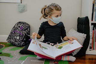 girl with mask on and backpack reading a book