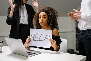 A woman at her work desk, surrounded by coworkers, holds up a small sign that reads “HELP.”