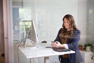 A woman is checking her phone, whilst at her desk with the computer in front of her. The impression is of someone who is both busy and happy in their workplace.