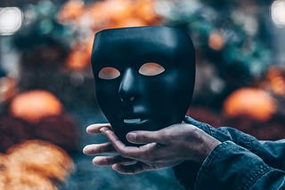 Person’s hands holding a black face mask over a blurred orange and teal background.