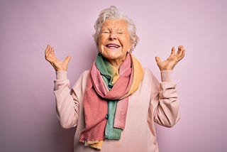 Senior lady on pink background, eyes closed with arms up in excitement