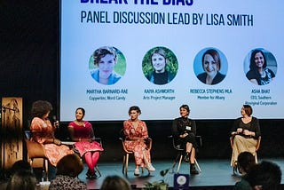 A diverse group of women sit on a stage in front of a screen. They’re participating in a panel discussion about bias on International Women’s Day 2022. Photo credit: Krysta Guille.