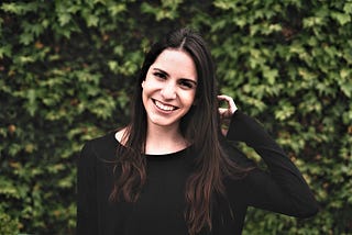 smiling girl with long dark hair wearing long sleeve black top standing in front of green landscape wall