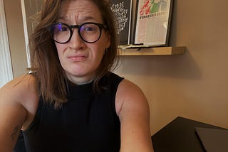 Selfie of me sitting at my desk with a frightened expression. I am wearing a sleeveless black high neck tank top, black thick glasses, and my hair is down and framing my face. I am in the centre of the photo but a little to the left and the corner of my desk is on the right. My desk is black, the wall behind me is brown.