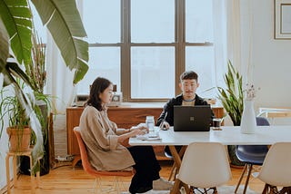 Two humans sitting at a white table with their computers out, surrounded by lush green plants and big windows in a lush office space.