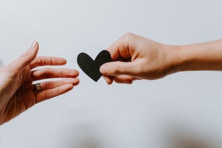 A pair of hands reaching out for each other. On the right, a light-skinned person is reaching out to hand a cutout black paper heart to another outstretched light-skinned hand on the left.