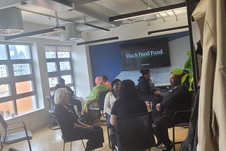 Image of a group of people witting in a room on chairs in a few small cirlces. There is a coat in the foreground and a plant visible. In the background is a screen with the words Black Food Fund. White writing. Black text.