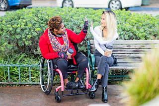 Person in wheelchair high-fiving person on park bench. From Getty Images.