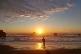 Wide angle photograph of a man from behind who’s facing the setting gold ball of the sun over Pacific ocean waves, with faint clouds overhead along the California coast.