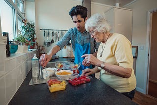 A young person with black short hair is standing in a kitchen with an elderly person with grey hair. The young person is wearing an apron, and helping serve fruit into bowls.