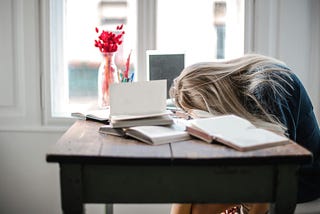 Photo of woman with her head down on wooden desk, long blond hair covering her face, surrounded by laptop, books, notebooks