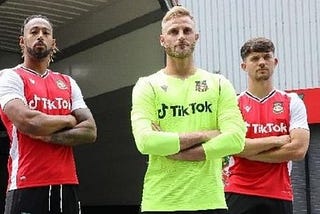 Three Wrexham FC players stand with arms crossed wearing their new jerseys with the TikTok logo emblazoned on the front