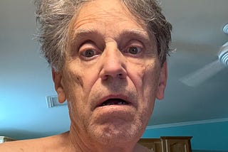 A man (me) with gray hair and a tired expression on his face, sitting in a room with light blue walls and a ceiling fan visible in the background.