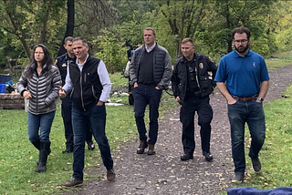 A picture of a group composed of Ithaca Common Council member Cynthia Brock, U.S. Congressperson Marc Molinaro, Tompkins County Legislator Mike Sigler, and Ithaca Police Department officers Thomas Condzella & John Joly walking down a wooded path. Some camping gear is seen at the edges of the image. Source: Local Electeds, Candidates Tour Homeless Encampment sites In Ithaca