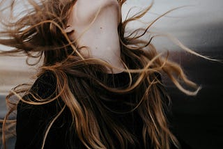 Close-up of a woman’s neck and long, wavy brown hair blowing wildly in the wind, with her face tilted upward and mostly out of frame, creating a sense of motion and intense emotion.