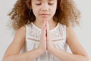 Photo of a young girl with curly hair, eyes closed and hands in “namaste” pose