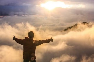 Silhouette of a young man seen from behind with arms spread, giving thumbs up as he floats in the sky amidst the clouds and the sun.