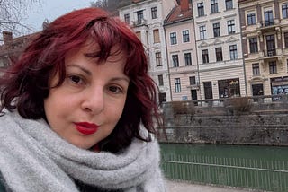 Sarah Jayne, a white woman with brightly dyed read hair looks into the camera. Behind here is a river and tall terraced houses in central Ljubljana.