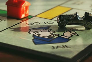 A close-up photo of a Monopoly game board, with a tiny car gamepiece resting on the “Go to Jail” space. A house gamepiece rests on the green property square next to it.