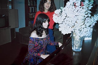 Two twenty-something sisters at a console piano, one playing and the other standing behind, singing, in a living room.