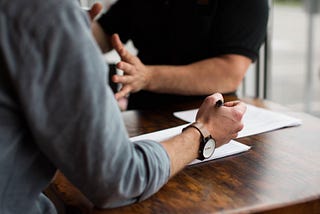 Two men talking at a conference table, while one man captures notes