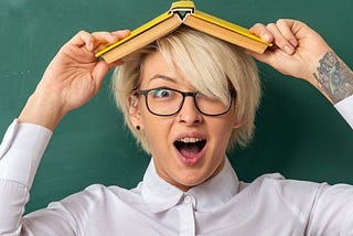 Excited young blonde female teacher wearing glasses in classroom standing in front of chalkboard holding book on head looking at camera