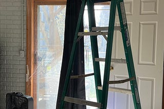 A view of a gray walled room with a ladder in its center sitting underneath a cut-out in a ceiling exposing duct work and register. A drop cloth is underneath the ladder and exposed ceiling full of tools, removed light fixture, and the excised drywall. Sunlight filters through the window in the background.