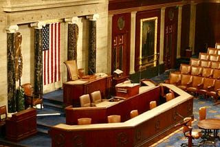 An interior view of the chamber of the US House of Representatives in the US Congress with an American flag displayed prominently at the front. The room features ornate woodwork, leather chairs arranged in rows, and a large desk with multiple chairs behind it. There is a portrait on the wall to the right and marble columns framing the flag. The carpet has a patterned design.