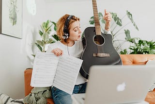 Woman Playing Guitar on Video Call on Computer. She is wearing a headset. She is pointing at the strings and has some sheet music in her right hand.