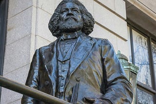 A photo of the Frederick Douglass Statue at the New York Historical Society, standing tall, proud and holding a book in his left hand close to his body.