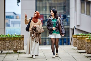 Two students walk across campus, smiling and pointing at buildings.
