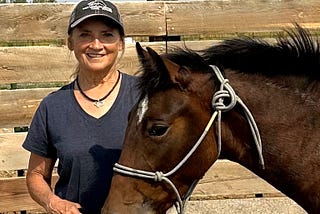 Whisper, a brown horse, and Nancy, a woman in jeans and a baseball cap, stand outside together in the sun.