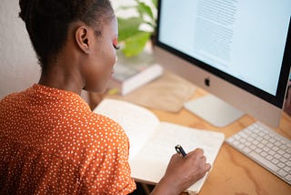 woman writing in notebook in front of computer screen, apple iMac, woman is writing with a pen, wearing an orange blouse and red make-up. Picture accompanies story by Katie on Medium about writing, publishing, and then deleting a story to then republish it.