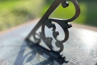 An aged bronze sundial casts a shadow on itself. The background is blurred, but appears to be grass and trees.