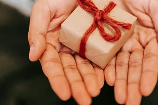 A pair of upturned hands holding a small brown parcel wrapped with red twine, in a gift offering gesture.